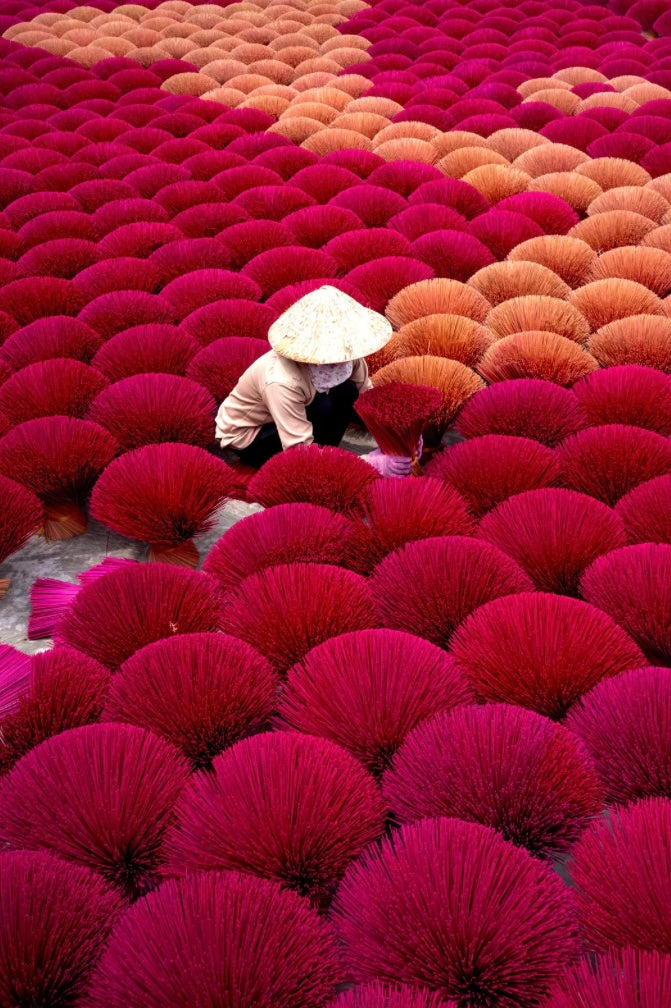 Artisan séchant des fagots d’encens colorés dans une cour, disposés en motifs circulaires rouges et ocres, créant un paysage vibrant et traditionnel d’Asie.